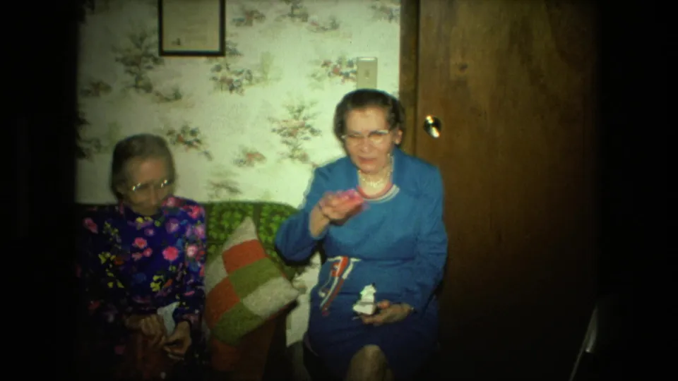 In a 1976 Fort Wayne home interior, two elderly women sit together as the woman in blue raises a small pink craft piece.