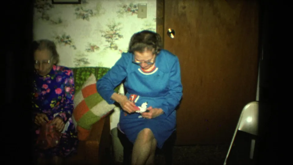 An elderly woman in blue concentrates on assembling or adjusting a small white craft object beside another woman on the couch.