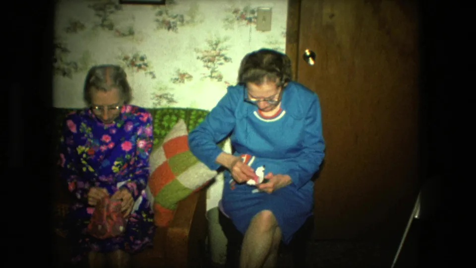 The 1976 home-movie frame shows two elderly women in a Fort Wayne living room during a quiet craft activity on the couch.