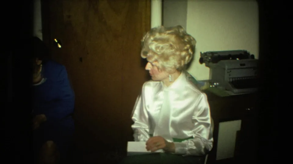 The woman in a white satin blouse turns toward another adult partly visible at left, with the envelope and typewriter still in view.