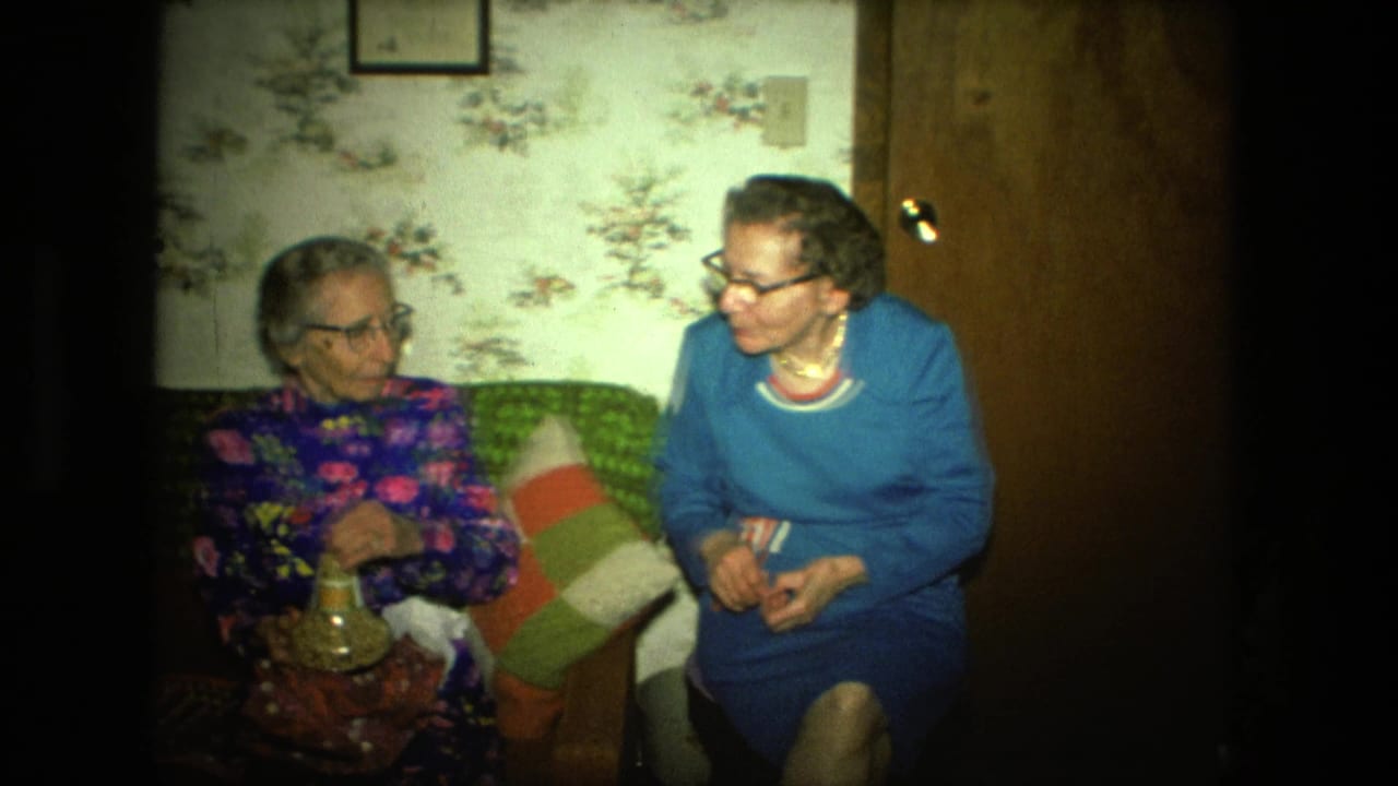 Two elderly women sit on a couch in a floral-wallpapered living room.