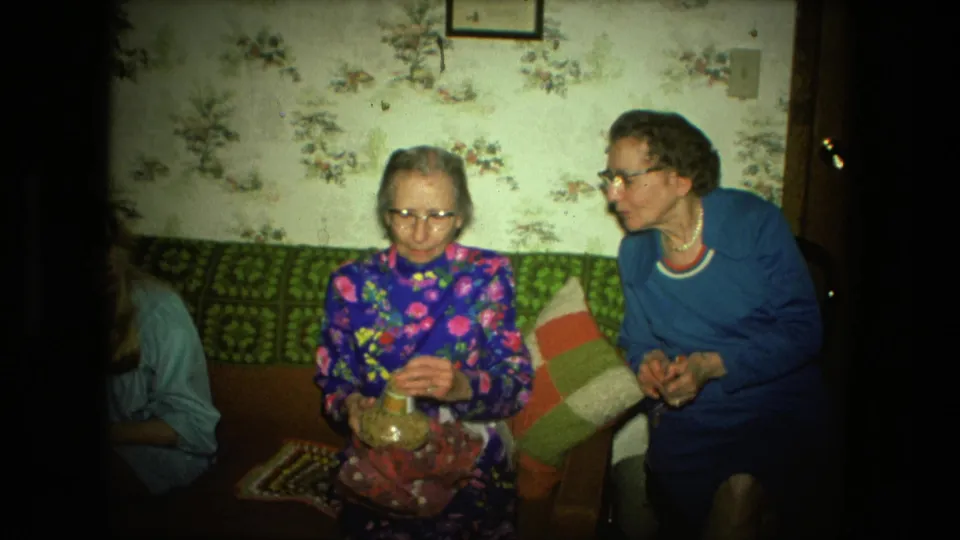 Two elderly women sit close on a green couch in a 1976 Fort Wayne home, with floral wallpaper and patchwork pillows behind them.