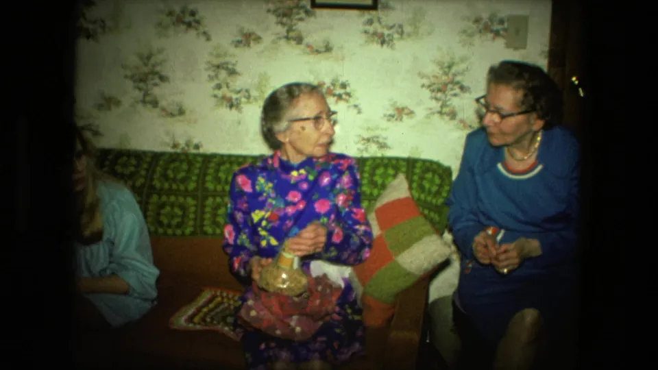 Two elderly women face each other on a couch in a Fort Wayne living room, with a glass jar, floral wallpaper, and pillows visible.