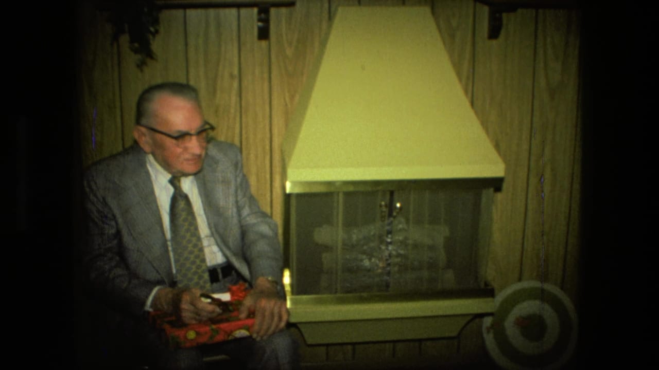 Older man in a gray suit sits beside a fireplace holding a red gift box.