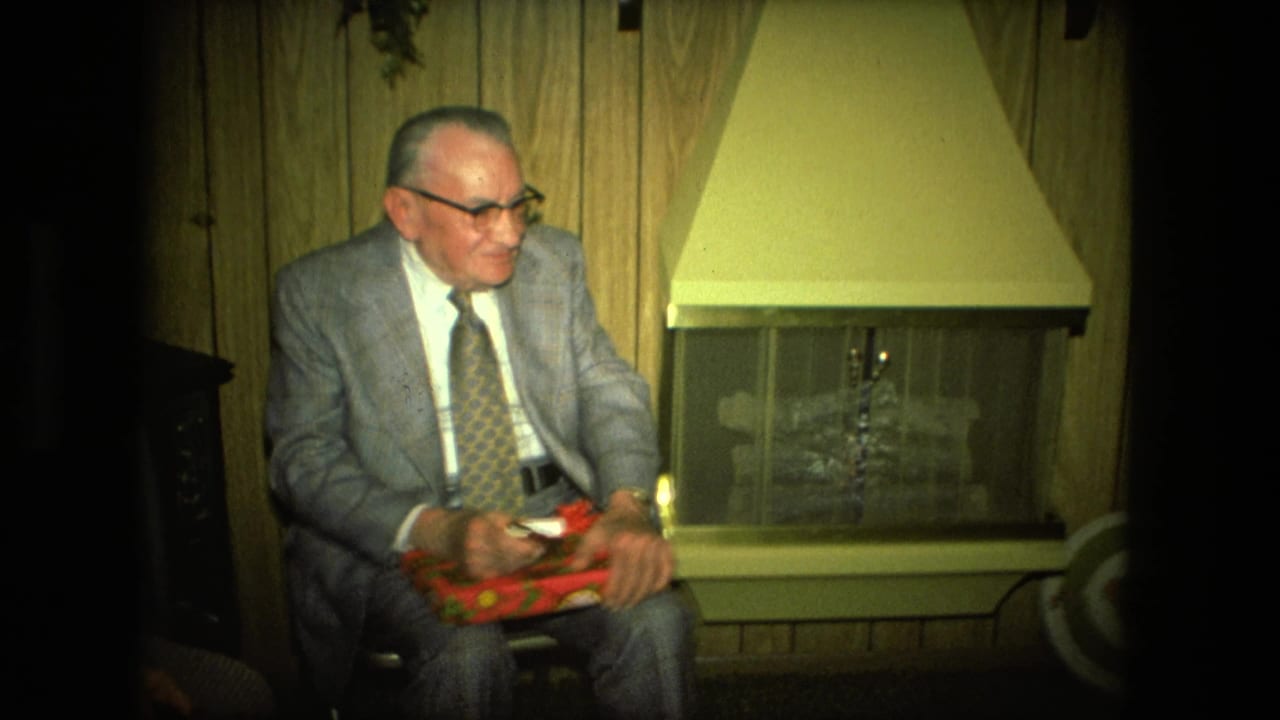 In a 1976 Fort Wayne living room, an older man in a suit handles a red wrapped gift by the fireplace.