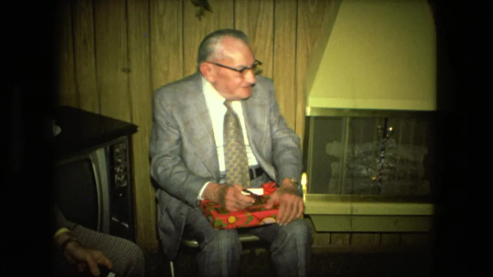 The 1976 home-movie frame shows a suited older man with a gift, a fireplace, and a vintage television in a private room.