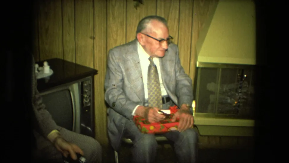 An older man in a gray suit sits by the fireplace with a partially opened red gift box in Fort Wayne in 1976.