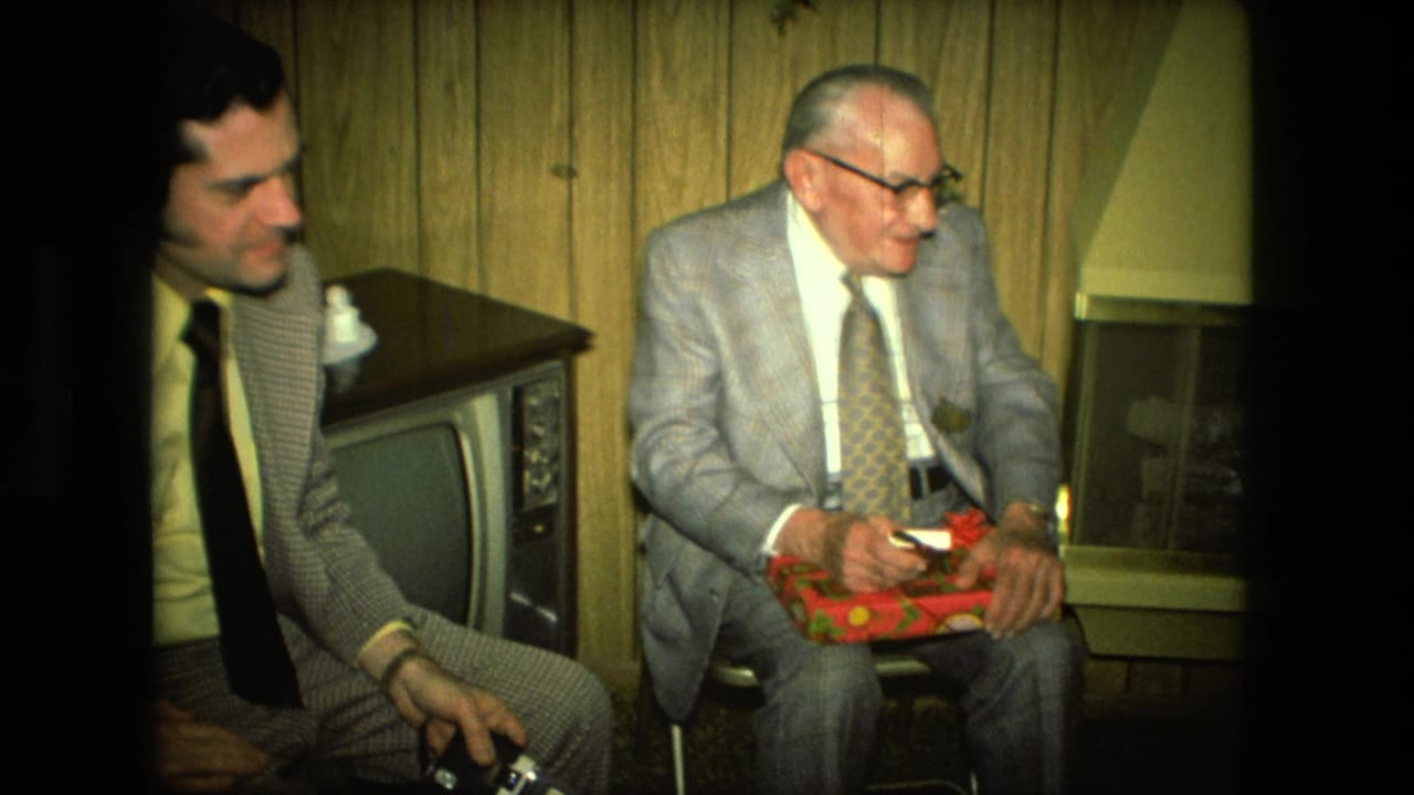 In a 1976 Fort Wayne home interior, two suited men sit near a vintage television while the older man holds a red gift box.