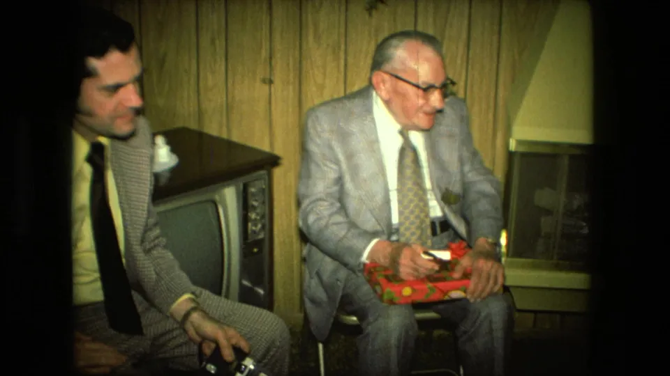 In a 1976 Fort Wayne home interior, two suited men sit near a vintage television while the older man holds a red gift box.