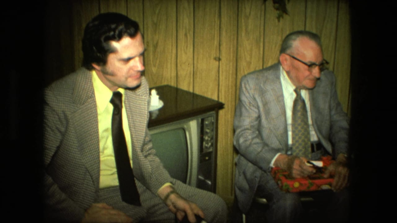 A younger man in a checkered suit sits beside an older man holding a red gift box.