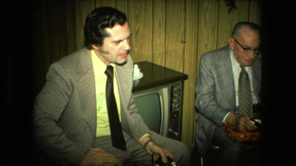 In the Fort Wayne living room, the younger man in a checkered suit is foregrounded as the older man sits behind him near the gift box.