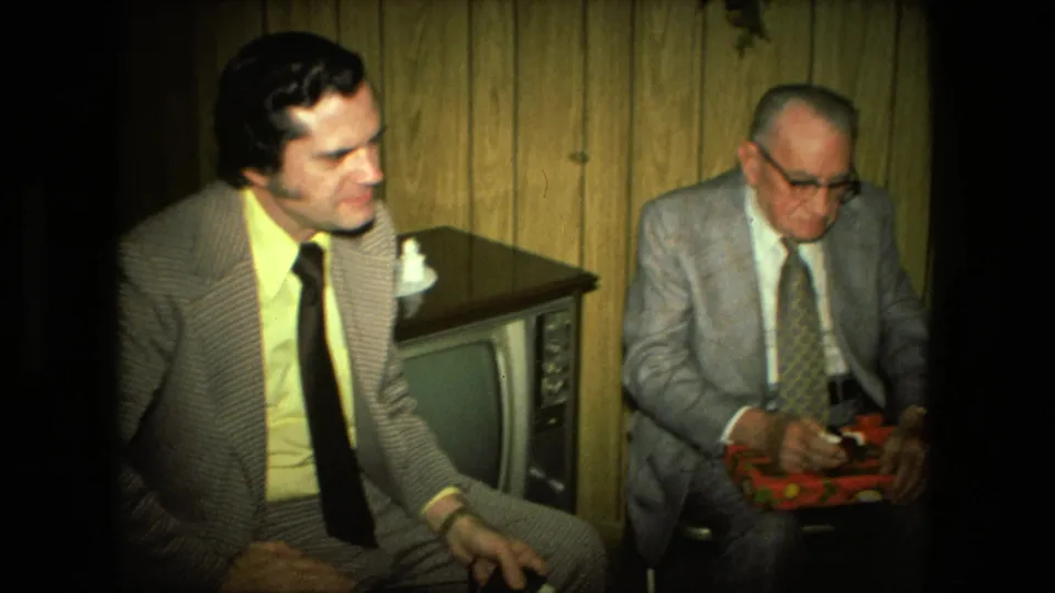 The 1976 home-movie frame shows two suited men seated near a television, with the older man looking down toward the red gift box.