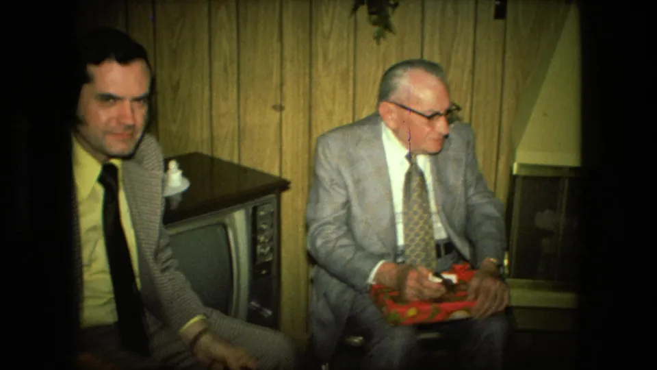In a 1976 Fort Wayne home-movie frame, two suited men sit in a wood-paneled living room near a console television.