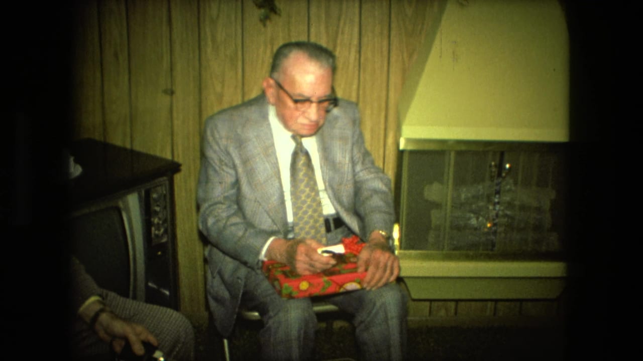 An older man in a suit sits by a fireplace holding a red wrapped gift.