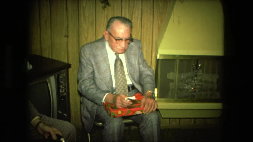 An older suited man sits beside a fireplace in a Fort Wayne living room, holding a red wrapped gift in 1976 home-movie footage.