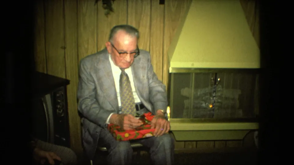 The older man remains seated near the fireplace, looking down at the red wrapped gift in a 1976 Fort Wayne interior scene.