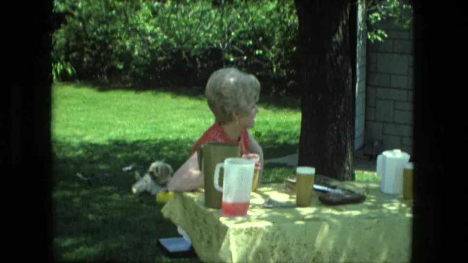 A woman sits at a backyard table with a drink pitcher and dishes, while a small dog rests on the grass in Fort Wayne home-movie footage.