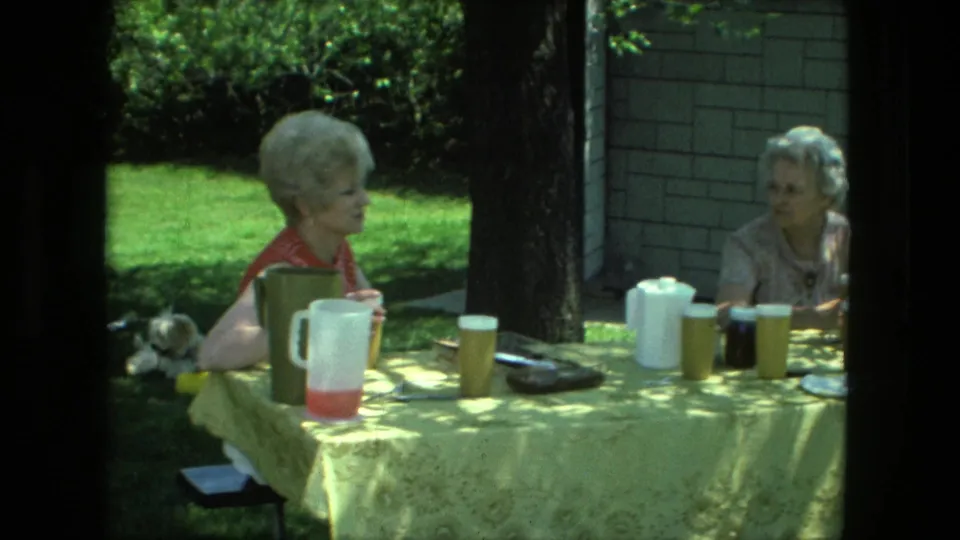 Two older women sit across a yellow-covered backyard picnic table in Fort Wayne, Indiana, with pitchers, cups, plates, and trees around them.