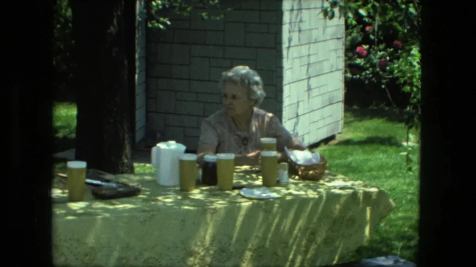 An older woman sits at a yellow-covered picnic table in a Fort Wayne backyard, with cups, pitchers, a dark jar, and food visible on the table.