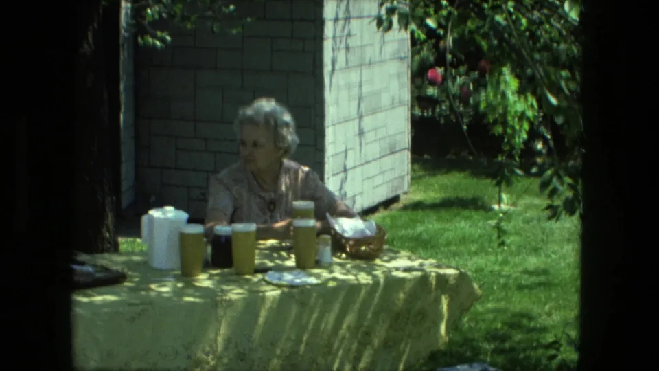 A Fort Wayne backyard frame shows an older woman seated at a picnic table with white pitchers, cups, a dark jar, and greenery in the background.