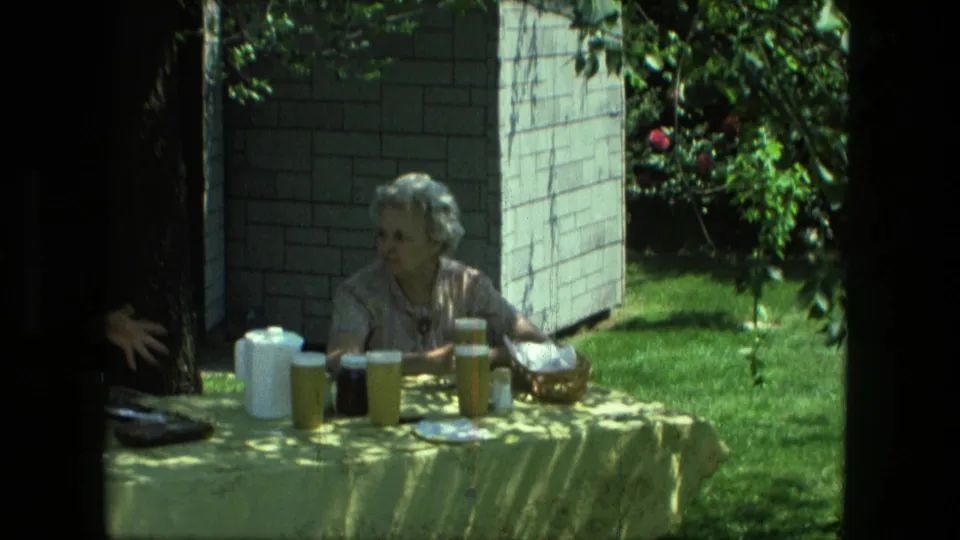 1976 Fort Wayne 8mm frame showing a woman at a shaded outdoor picnic table with drink pitchers and cups.