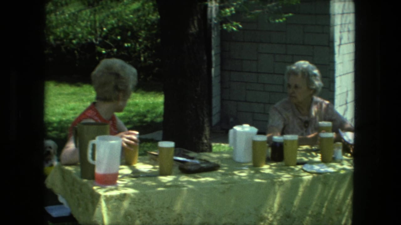 Two women sit at an outdoor picnic table with pitchers, cups, and dappled sunlight.