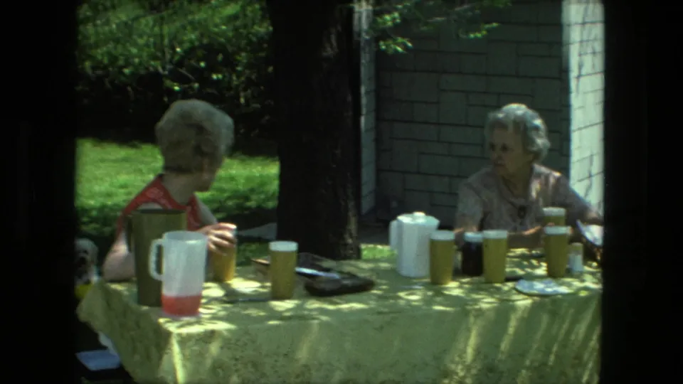 1976 Fort Wayne home-movie frame of two women at a yellow-covered picnic table in a shaded yard.