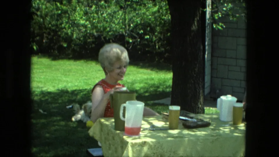 1976 Fort Wayne home-movie frame of a woman smiling at a shaded outdoor table with pitchers and cups.