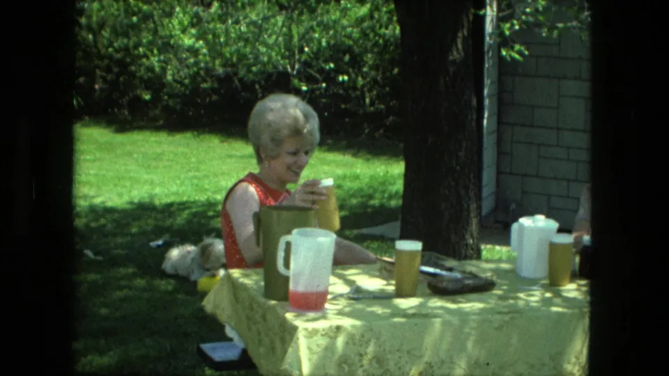 In 1976 Fort Wayne home-movie footage, a woman sits at a yellow-covered backyard table with pitchers and cups as a small dog lies on the grass.