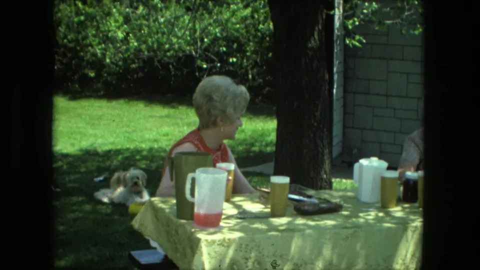 The woman turns away from the camera at a Fort Wayne backyard table, with pitchers, paper cups, shade trees, and a small dog visible behind her.