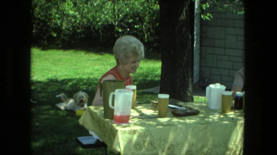 A woman in a red sleeveless dress looks down beside a pitcher and cups on a yellow tablecloth in a 1976 Fort Wayne backyard scene.