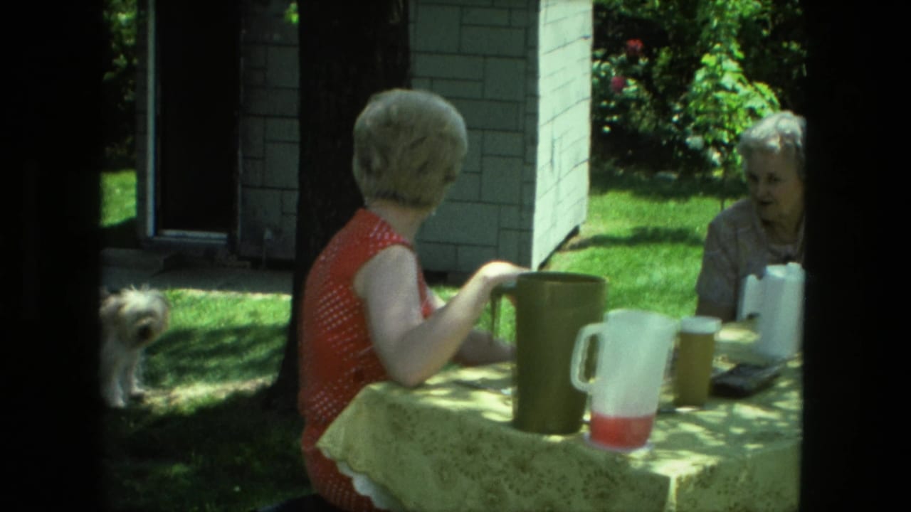 Two women sit at a shaded outdoor table with pitchers and cups visible.
