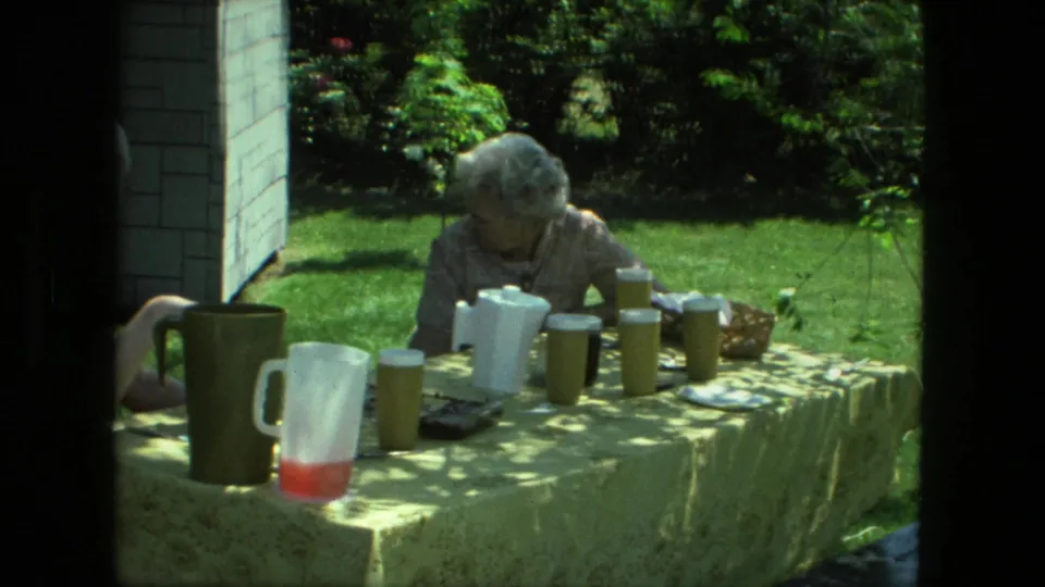 1976 Fort Wayne backyard meal frame with a woman at the far side of a table covered in yellow cloth, pitchers, cups, and plates.