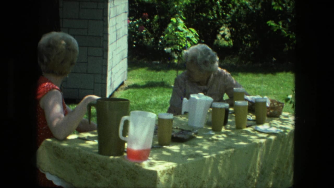 Two women sit outdoors at a table with pitchers and cups in a shaded yard.