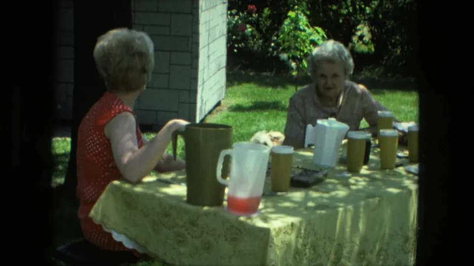 1976 Fort Wayne 8mm frame of two women at a backyard table, with the older woman facing camera and refreshments between them.