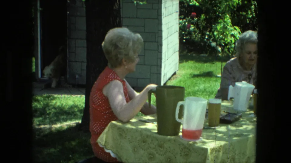 A smiling woman in a red polka-dot dress sits at a yellow-covered table with pitchers as another woman is visible across the table.