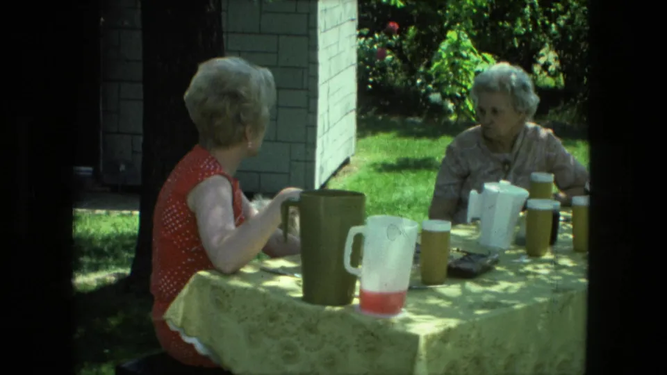 1976 Fort Wayne frame showing two women socializing at an outdoor table with drink pitchers, cups, and a patterned tablecloth.