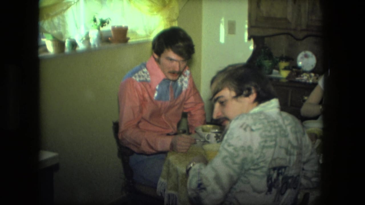 1976 Fort Wayne 8mm frame showing two seated men at a lunch table in a private kitchen with dishes and a wood cabinet.