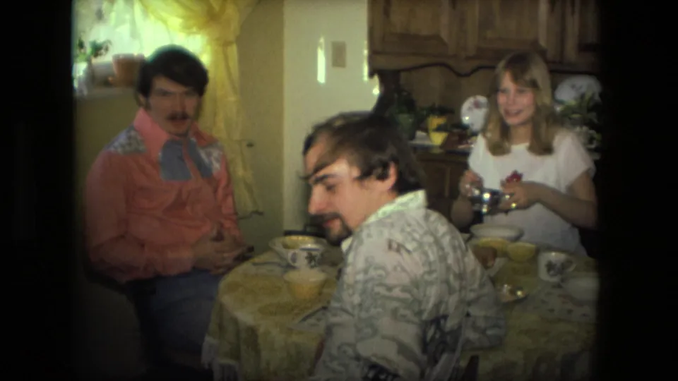 1976 Fort Wayne kitchen lunch frame with a man looking back toward the camera and a woman holding tableware beside him.