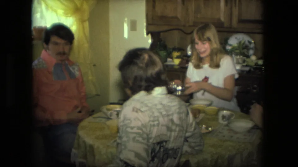 1976 Fort Wayne home-movie frame of a smiling woman and two seated men at a lunch table in a private kitchen.