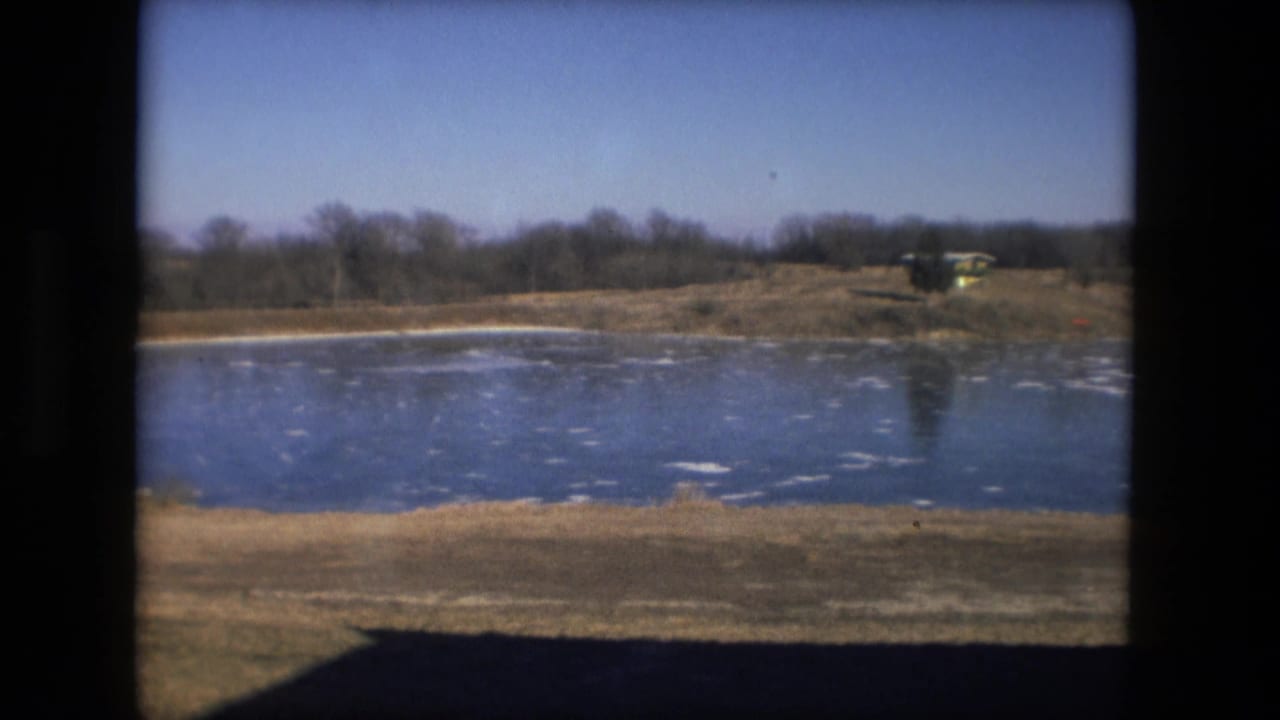 Frozen pond with scattered ice patches, brown grass, bare trees, and distant low buildings.