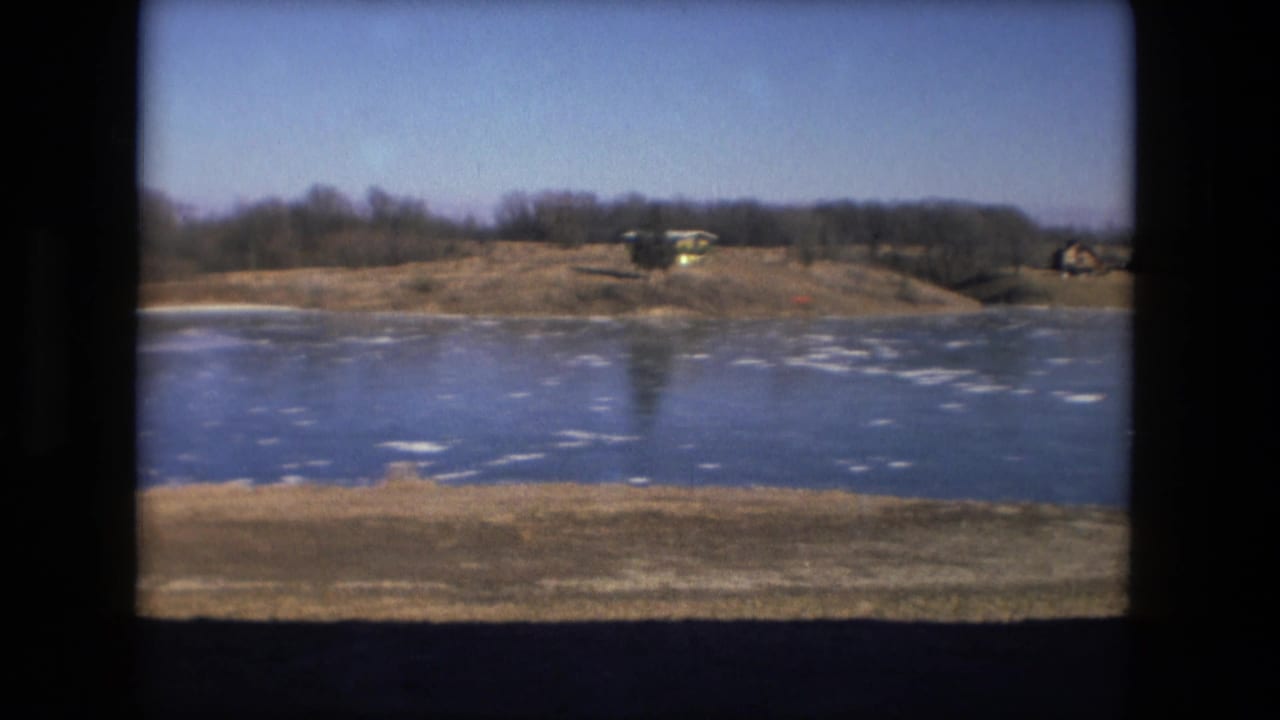 Fort Wayne winter pond scene on 8mm film, with ice patches across the water and low structures beyond the shoreline.