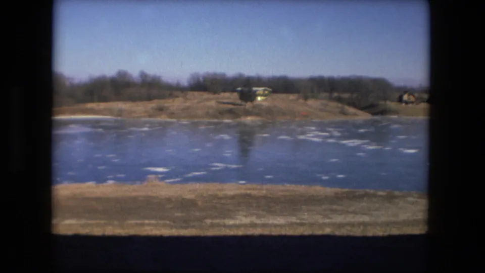 Fort Wayne winter pond scene on 8mm film, with ice patches across the water and low structures beyond the shoreline.