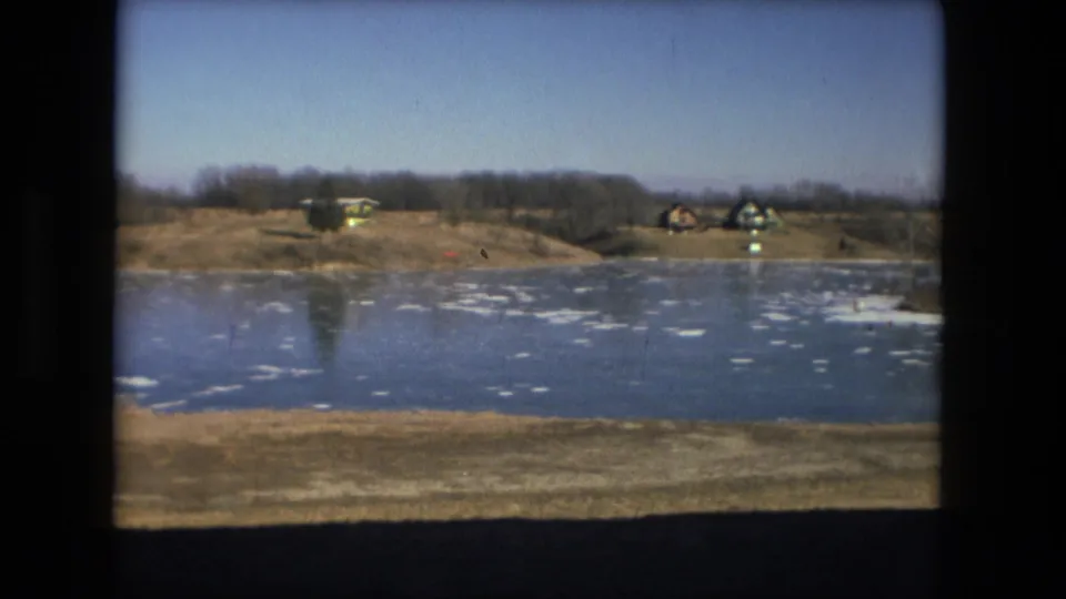 1976 Fort Wayne home-movie frame of a frozen pond and far bank with small buildings, bare trees, and winter grass.