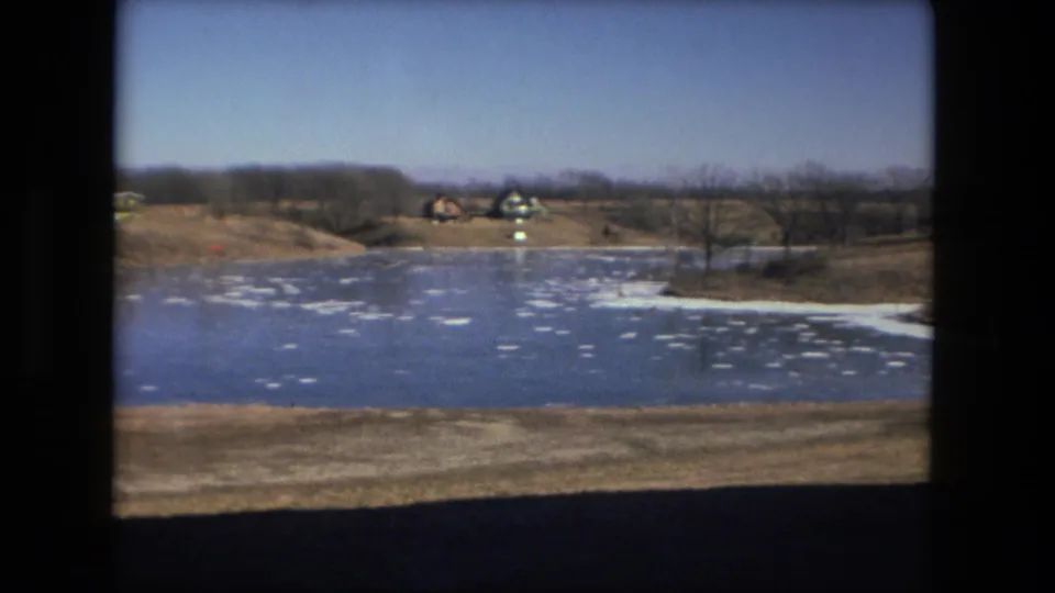 Fort Wayne 8mm winter frame showing an ice-covered pond, distant shoreline structures, leafless trees, and a clear sky.