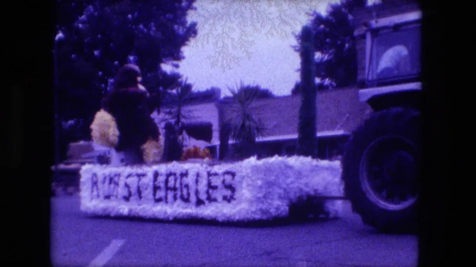 A floral parade float with visible lettering passes through Fort Wayne in 1976, with a tractor partly visible at the right edge.