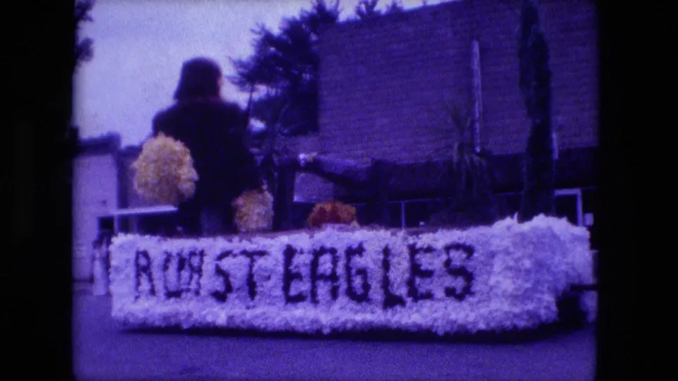 Close 1976 Fort Wayne frame of the floral float with dark lettering, a figure or decoration above it, and a brick building behind.