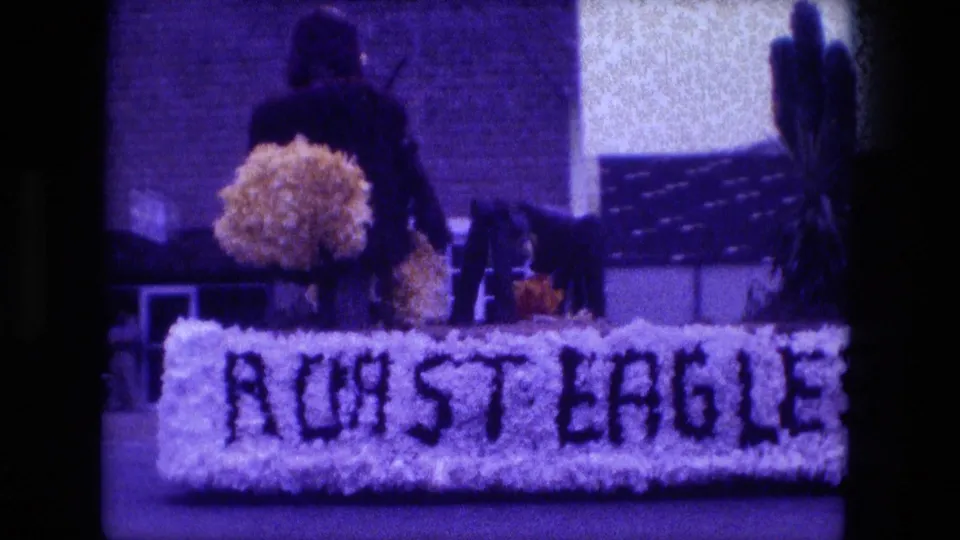 Close 1976 parade view of the Roast Eagle text on the floral float, with a person partly visible behind the display.
