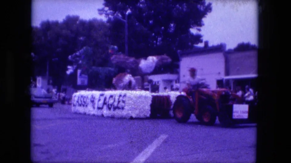 Wide Fort Wayne parade view with a red tractor pulling the Roast Eagles float along a tree-lined street.