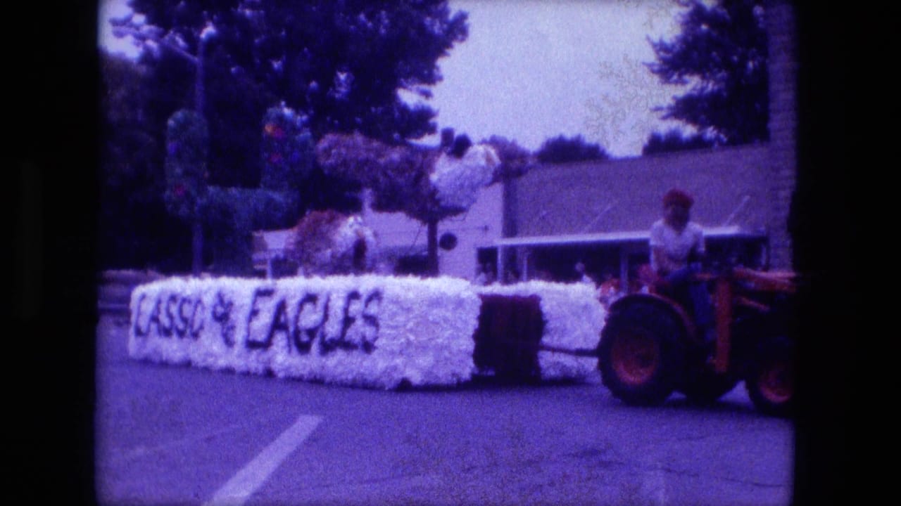 White EAGLES float moving behind a red tractor, with greenery and buildings behind it.